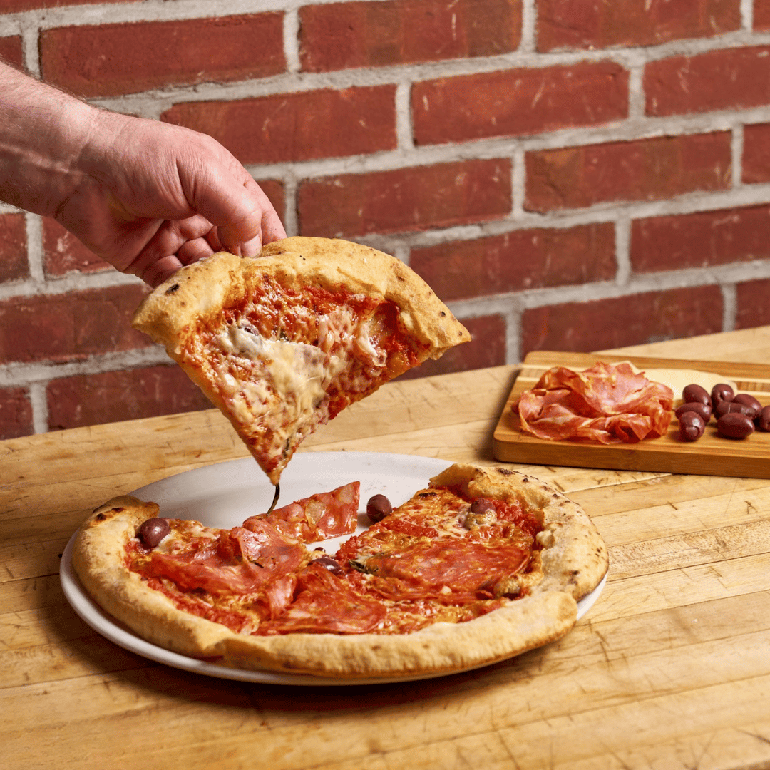 Slice of gluten free Padrino pizza with sopressata and olives being lifted, olives and salami on a board behind